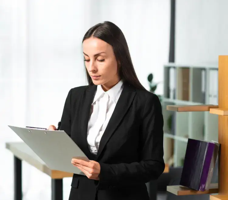 A professional woman in a business suit reviewing a clipboard in a well-lit office space.