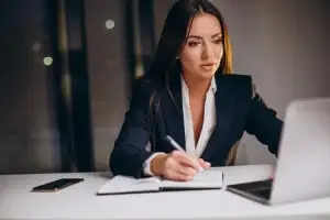 A professional woman in a suit writing notes while working on a laptop at her desk, with a smartphone beside her.