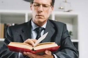 A mature male professional in a suit reading a red legal book in an office setting.